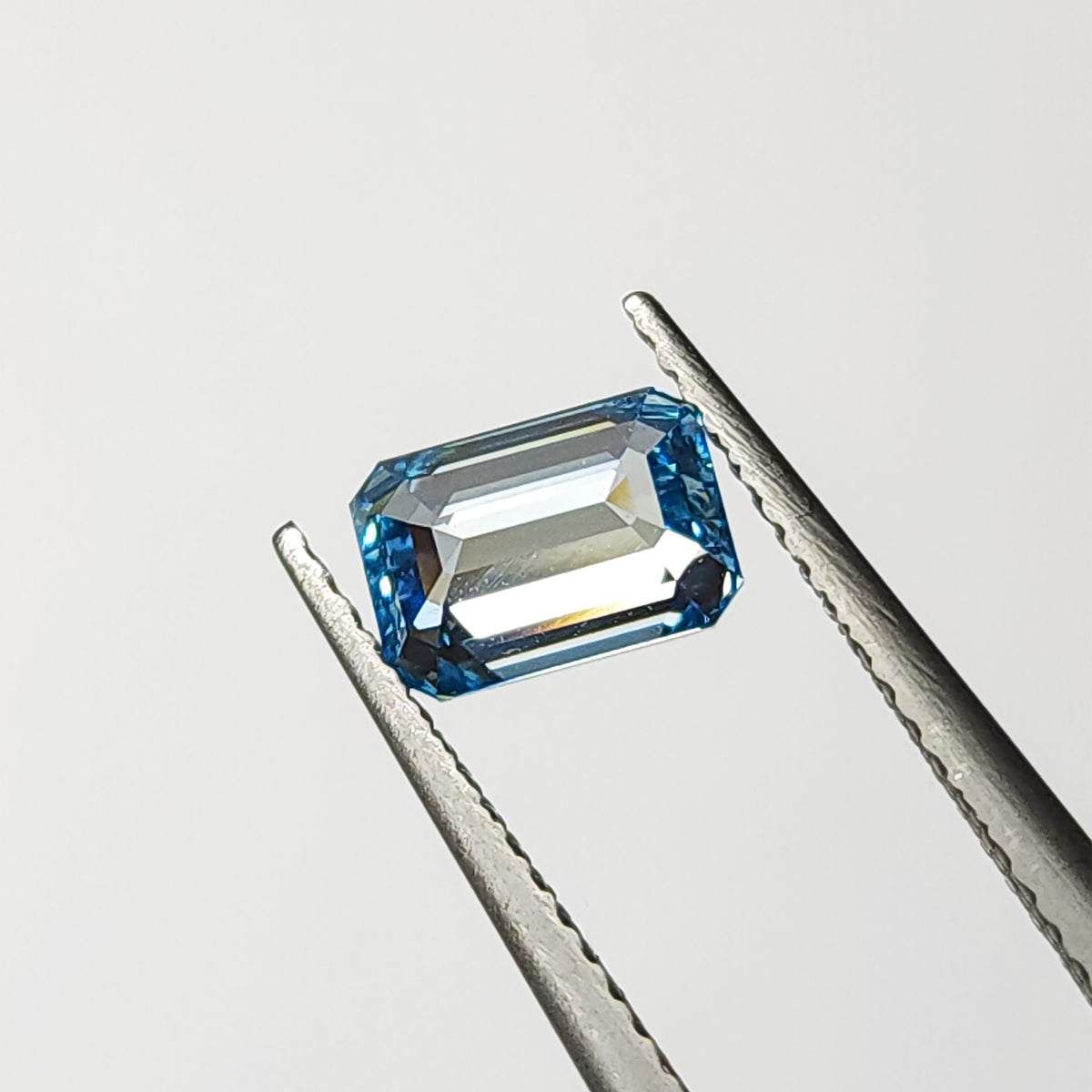Emerald-cut blue lab-grown diamond held face-up horizontally in tweezers with bright table reflections.