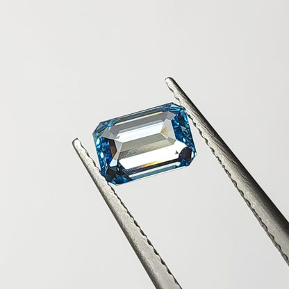 Emerald-cut blue lab-grown diamond held face-up horizontally in tweezers with bright table reflections.