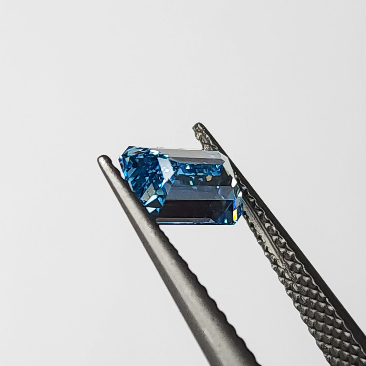 Side-profile view of the blue emerald-cut lab-grown diamond in tweezers, showing crown and pavilion depth.