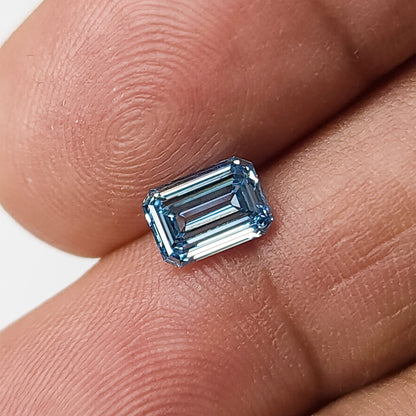 Blue emerald-cut lab-grown diamond resting face-up on a fingertip for scale.