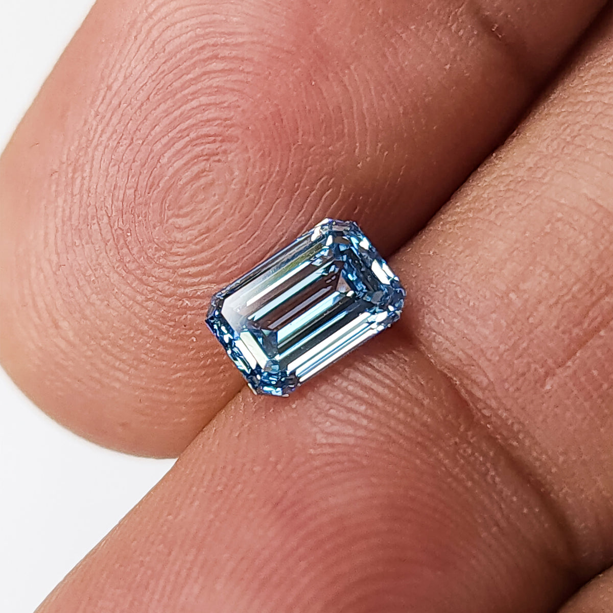 Emerald-cut blue lab-grown diamond lying face-up across fingertips from an alternate angle.
