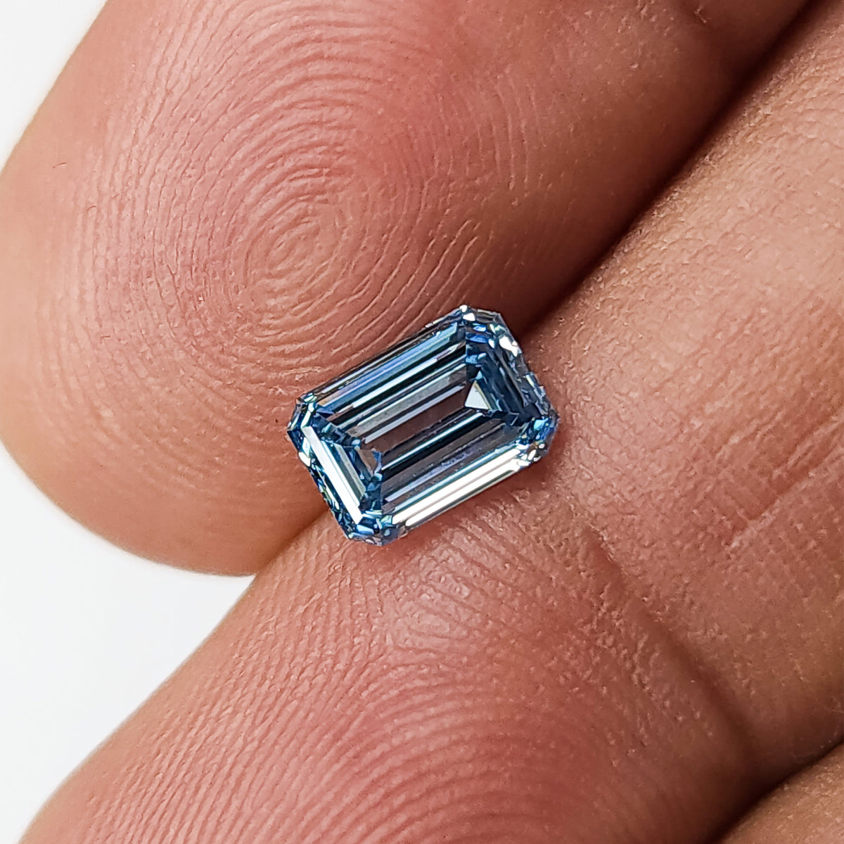 Close face-up view of the rectangular blue lab-grown diamond on fingertips, showing clipped corners.