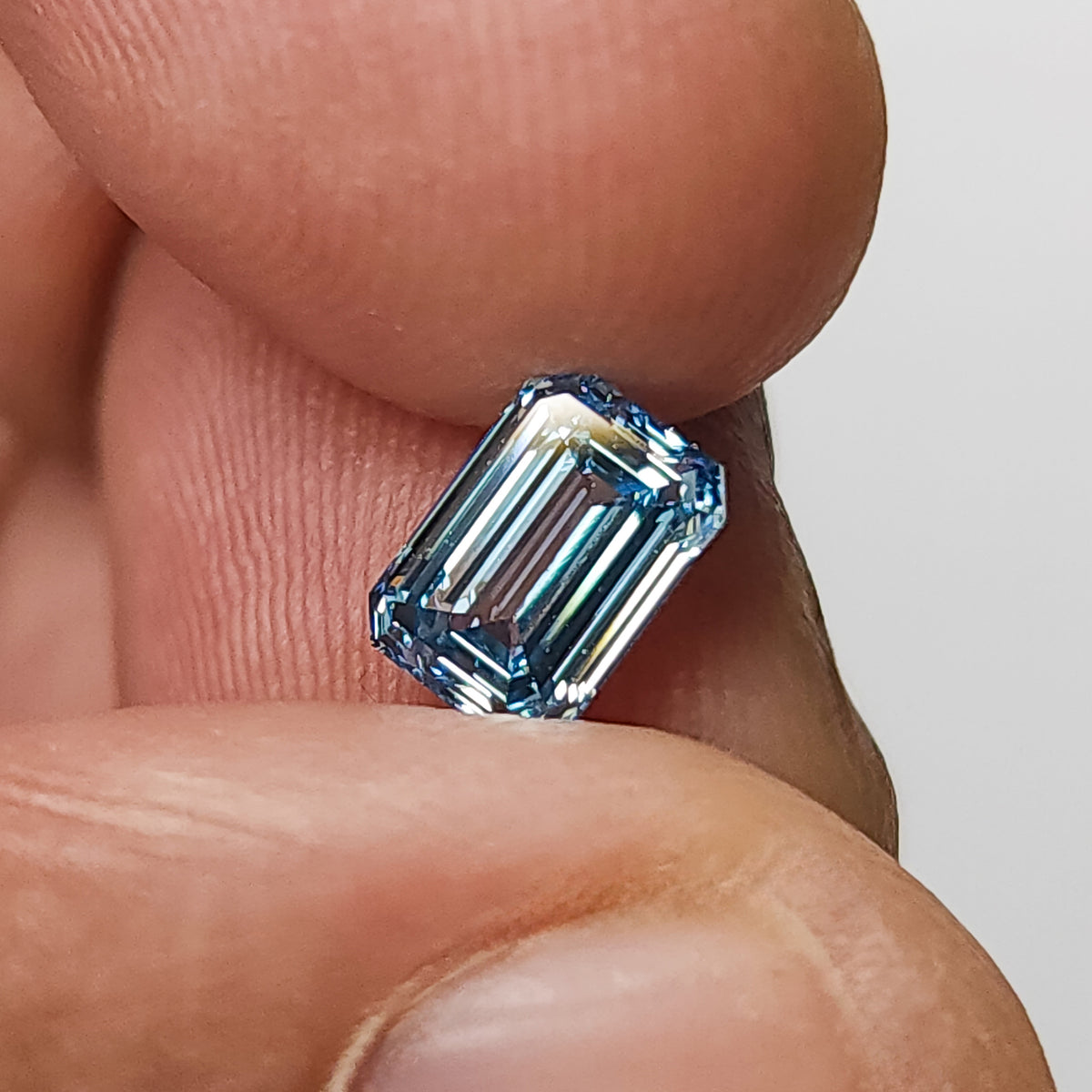 Blue emerald-cut lab-grown diamond held upright between fingertips for scale.
