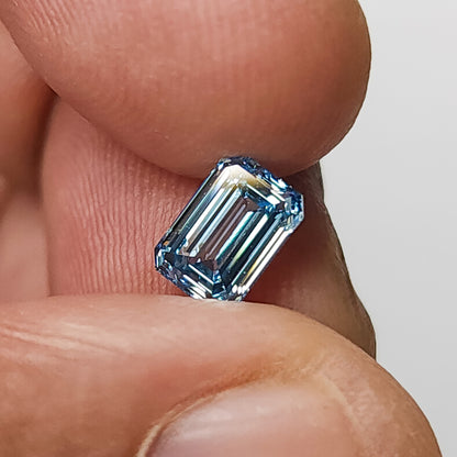 Blue emerald-cut lab-grown diamond held upright between fingertips for scale.