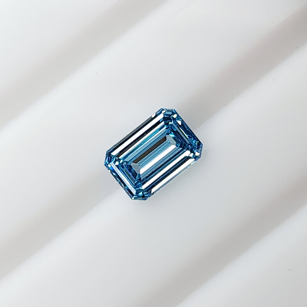 Blue rectangular emerald-cut lab-grown diamond lying face-up on a white grooved surface.