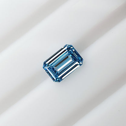 Blue rectangular emerald-cut lab-grown diamond lying face-up on a white grooved surface.