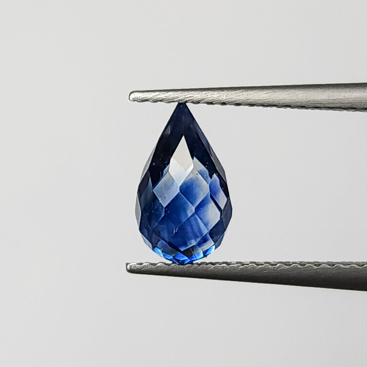 Briolette de zafiro facetado azul real sostenida con la punta hacia arriba con pinzas, mostrando su contorno completo de gota sin taladrar y reflejos brillantes de facetas.