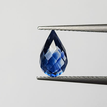 Briolette de zafiro facetado azul real sostenida con la punta hacia arriba con pinzas, mostrando su contorno completo de gota sin taladrar y reflejos brillantes de facetas.