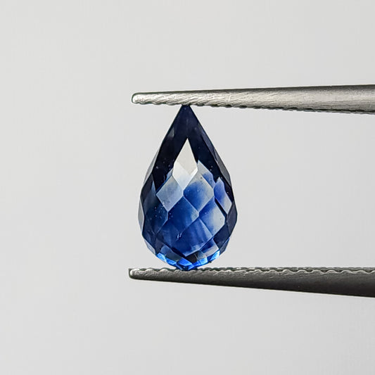 Royal-blue faceted sapphire briolette held point-up in tweezers, showing its full undrilled drop outline and bright facet reflections.