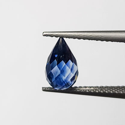 Briolette de zafiro azul sostenida verticalmente en pinzas desde un ángulo ligeramente girado, con la forma de gota completa visible.