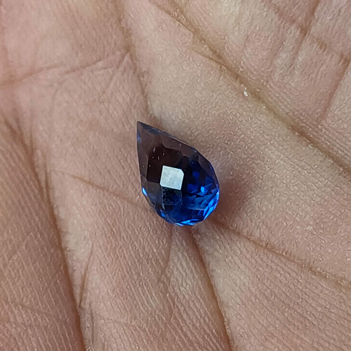 Briolette de zafiro azul facetado descansando sobre una palma para escala, mostrando su contorno de gota puntiaguda y reflejos.