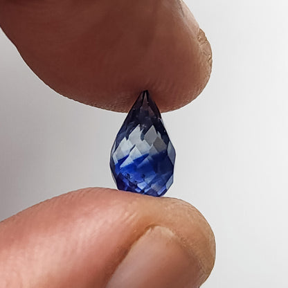 Briolette facetada azul equilibrada entre las puntas de los dedos para escala, con su forma de gota completa sin taladrar visible.