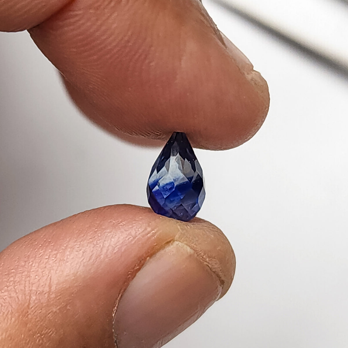 Briolette de zafiro facetada sostenida entre las puntas de los dedos desde otro ángulo, mostrando reflejos de facetas azules y pálidas.