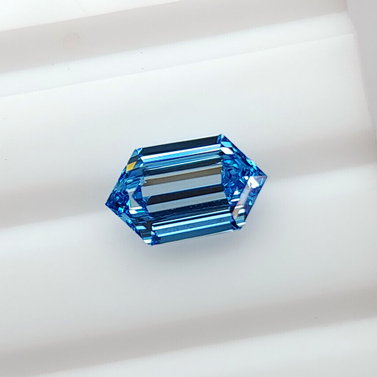 Blue elongated hexagon step-cut diamond viewed face-up on a white surface, showing parallel reflections across the center.