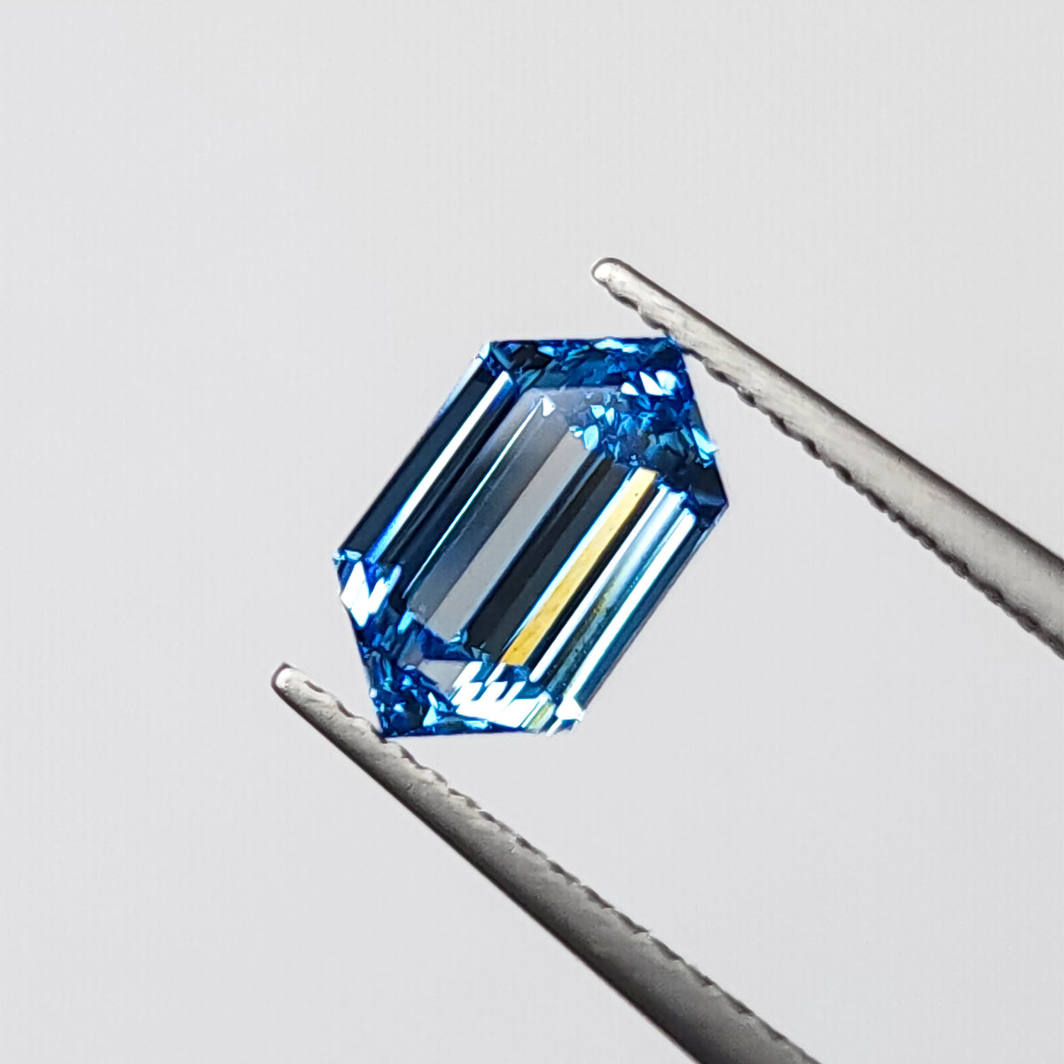 Blue elongated hexagonal diamond held face-up in tweezers, showing its six-sided outline and step facets.