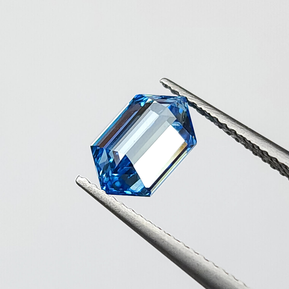 Blue hexagonal diamond tilted in tweezers, showing broad pale reflections inside the outline.