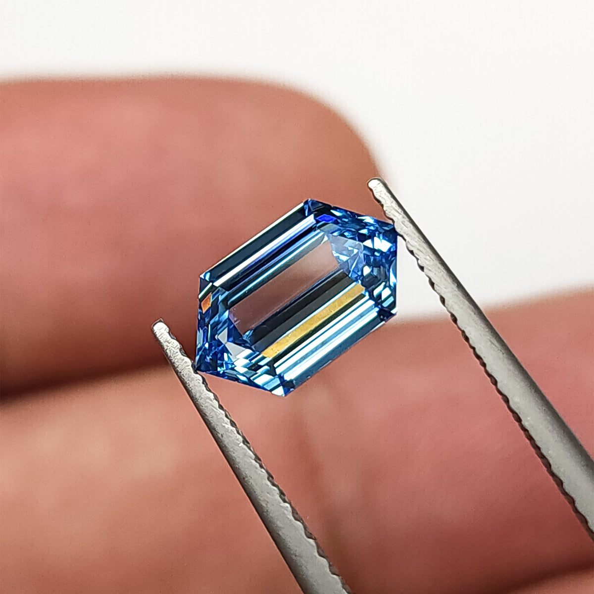 Blue step-cut hexagonal diamond in tweezers with a finger behind it, giving scale and showing darker center reflections.
