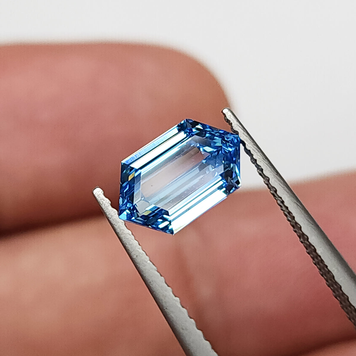 Alternate tweezers view of the blue hexagonal diamond near a fingertip, with bright pale reflections across the center.