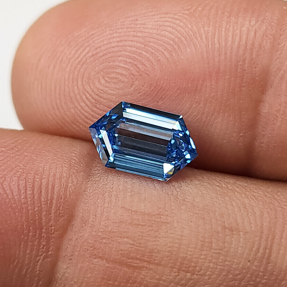Blue elongated hexagon diamond resting face-up on a fingertip, showing its size against fingerprint ridges.