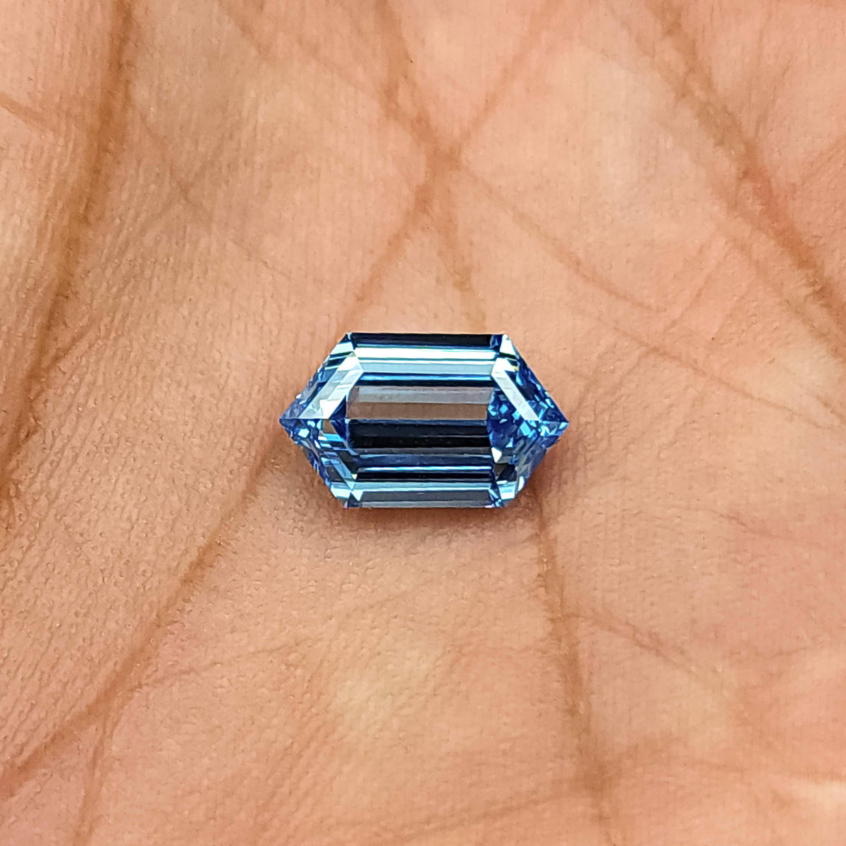 Blue hexagonal step-cut diamond viewed face-up in a palm, showing its outline and darker center bands.