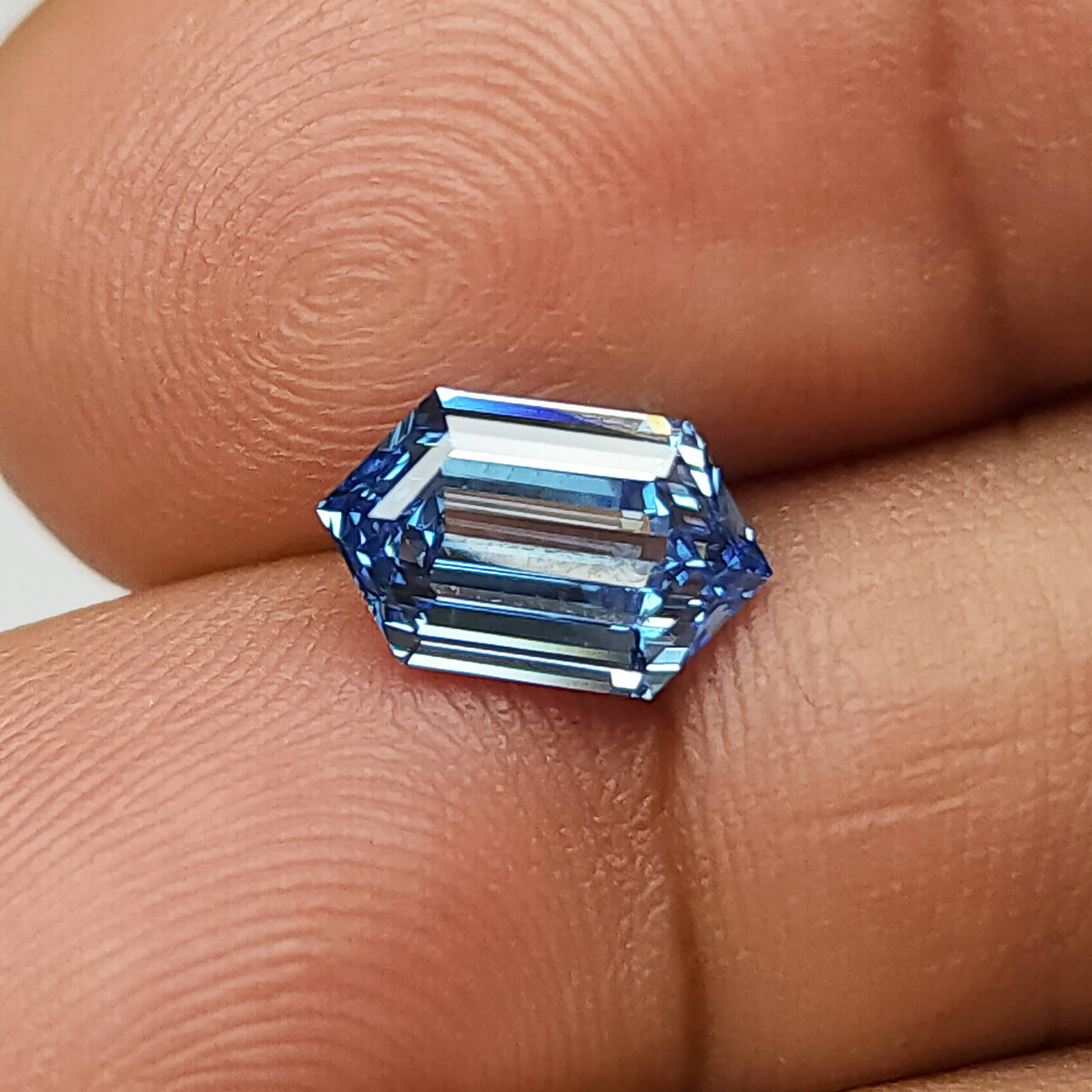 Alternate fingertip view of the blue elongated hexagonal diamond, with bright linear facet reflections.