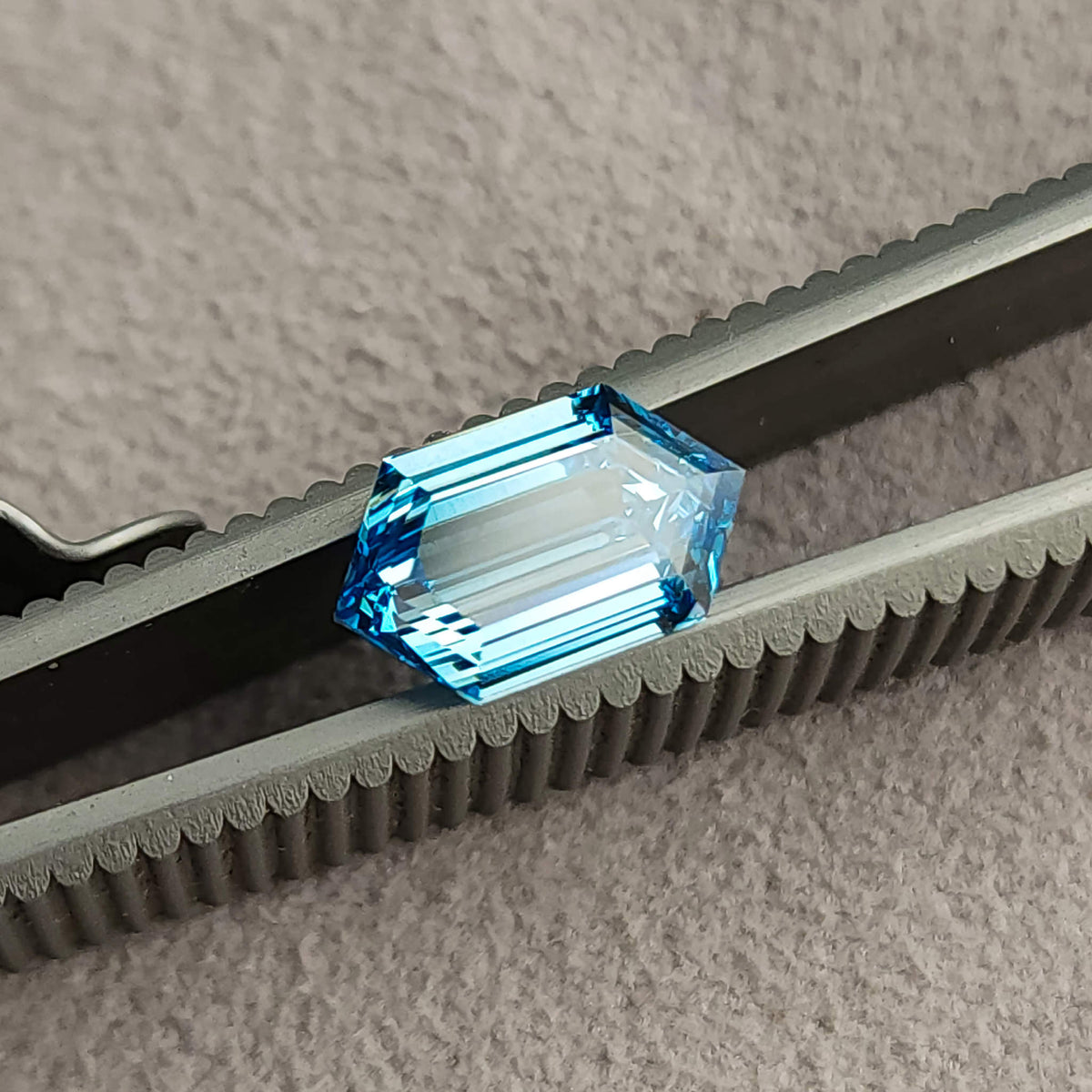 Oblique close-up of the blue hexagonal diamond held between tweezers against a gray surface.