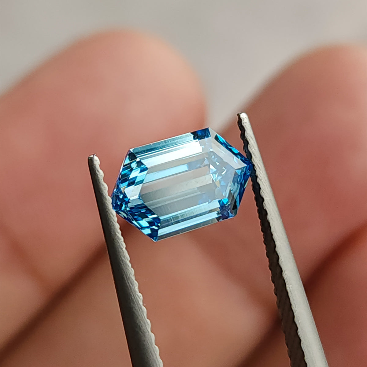 Blue step-cut hexagonal diamond in tweezers near a fingertip, with broad bright center reflections.