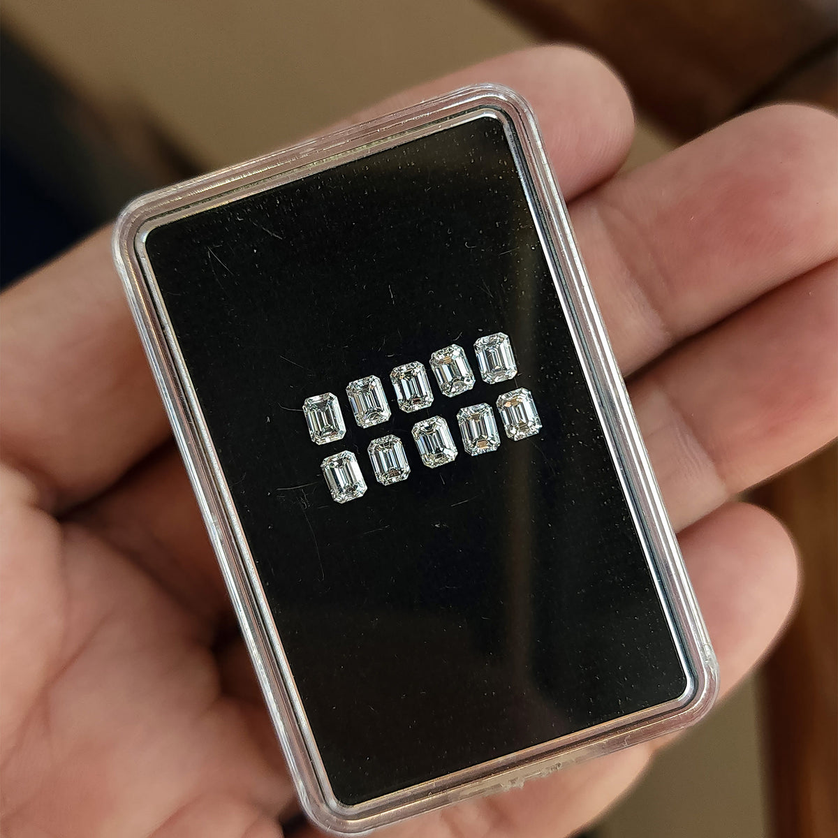 Hand holding a black display box with two rows of representative emerald-cut diamond stock.