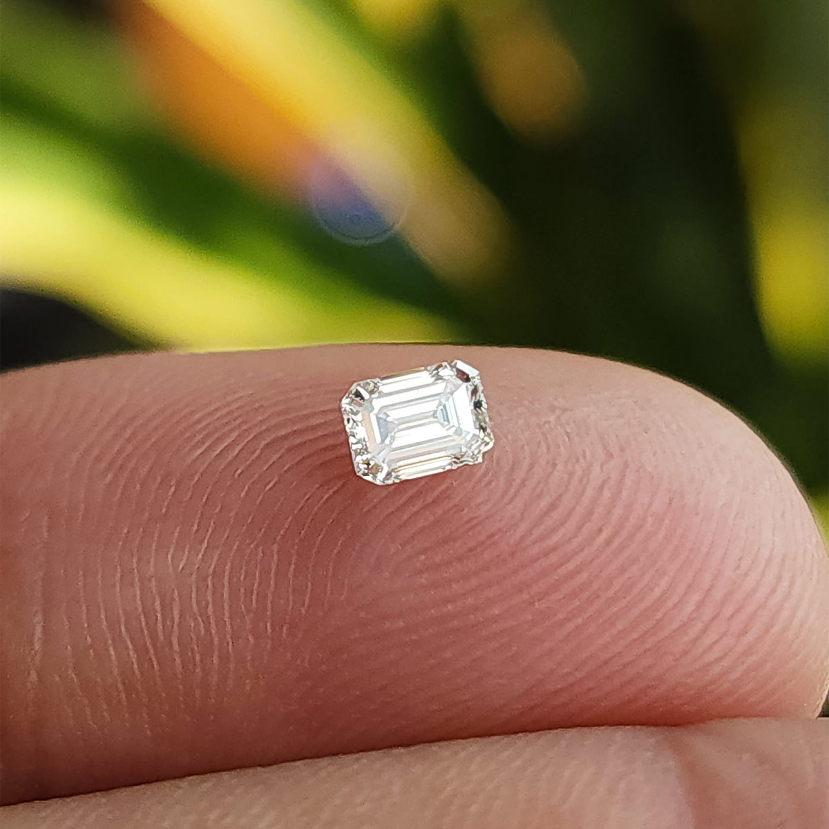Single emerald-cut diamond resting crown-up on a fingertip for scale against a green background.