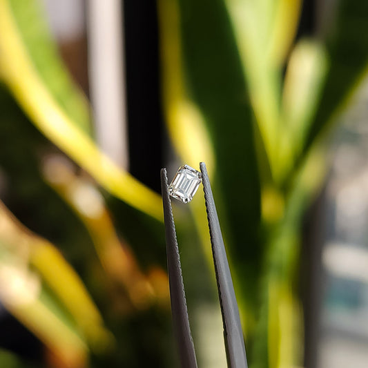 Single natural emerald-cut diamond held crown-up in tweezers, showing its rectangular table, clipped corners and step facets.