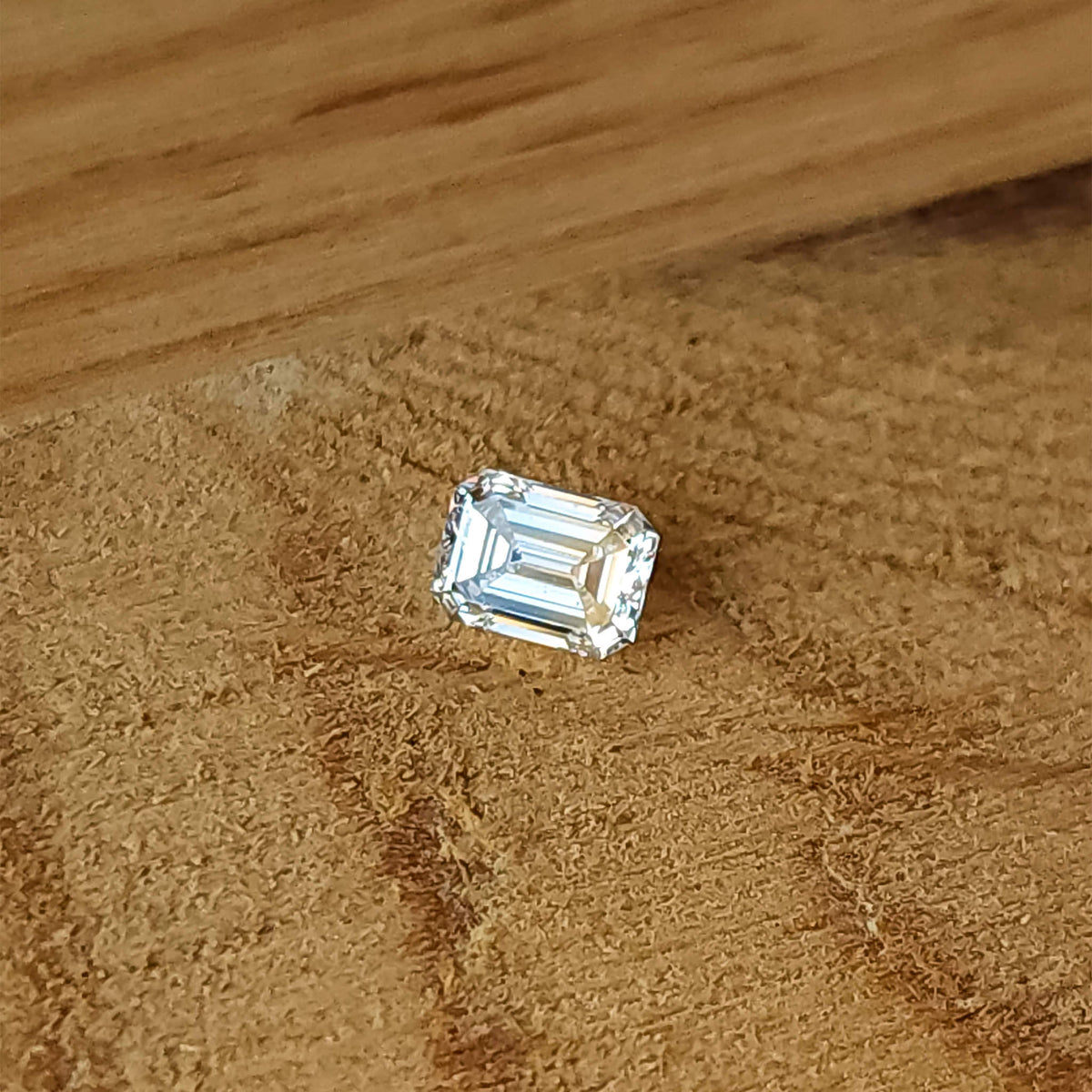 Single rectangular emerald-cut diamond lying crown-up on a wooden surface, with its step pattern visible.