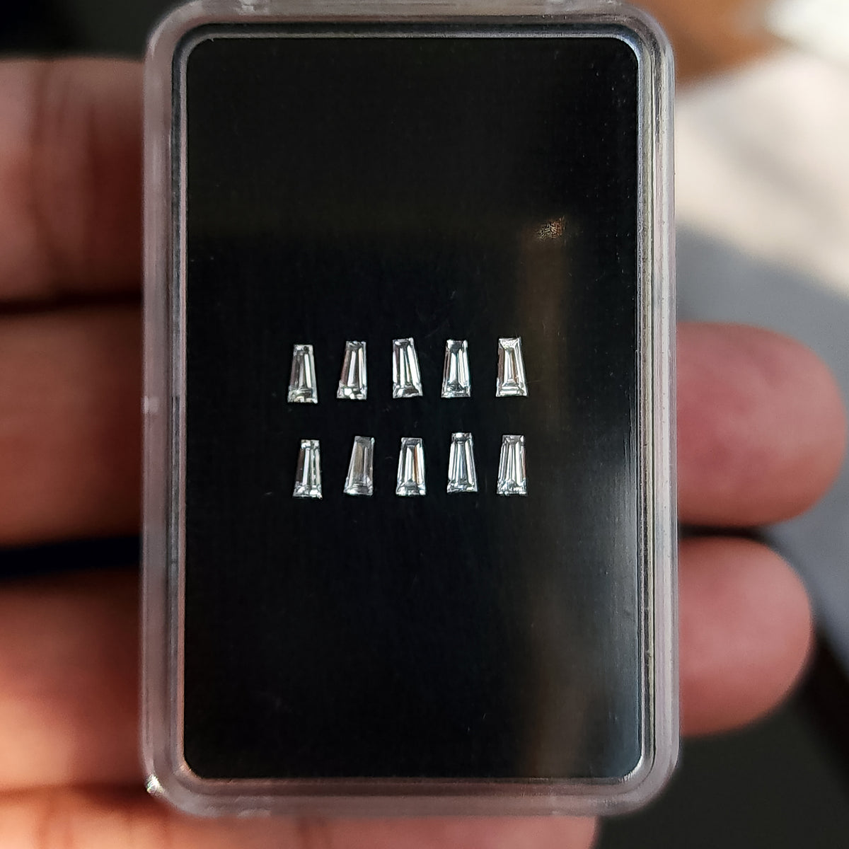 Diamantes baguette trapezoidales sobre una almohadilla negra en una caja transparente sostenida por una mano.