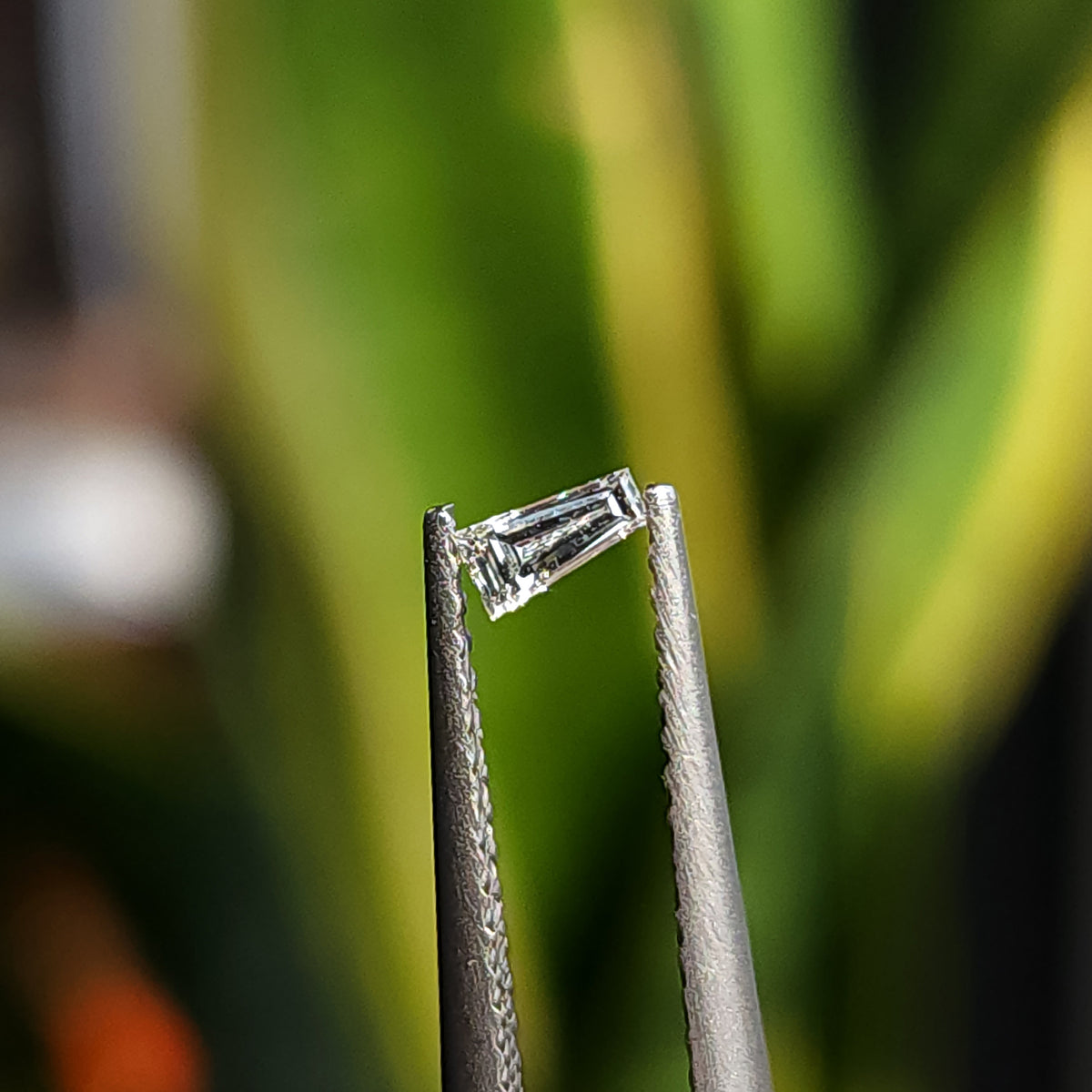 Un diamante natural baguette trapezoidal sostenido en pinzas sobre un fondo verde borroso.