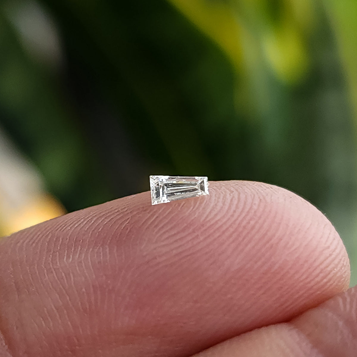 Un diamante baguette trapezoidal transparente descansando sobre la punta de un dedo para escala relativa.