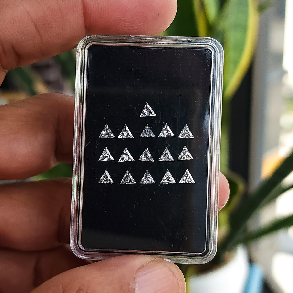 Black box of calibrated triangle diamonds held upright between fingers, with surrounding skin and a soft background for scale.