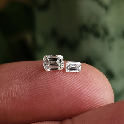 Two emerald-cut diamonds of different displayed sizes resting crown-up on a fingertip for comparison.
