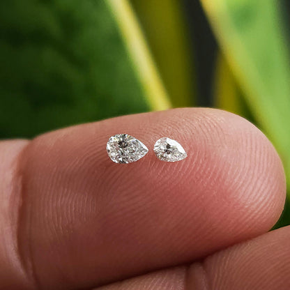 Two pear brilliant diamonds of different displayed sizes resting crown-up on a fingertip for comparison.