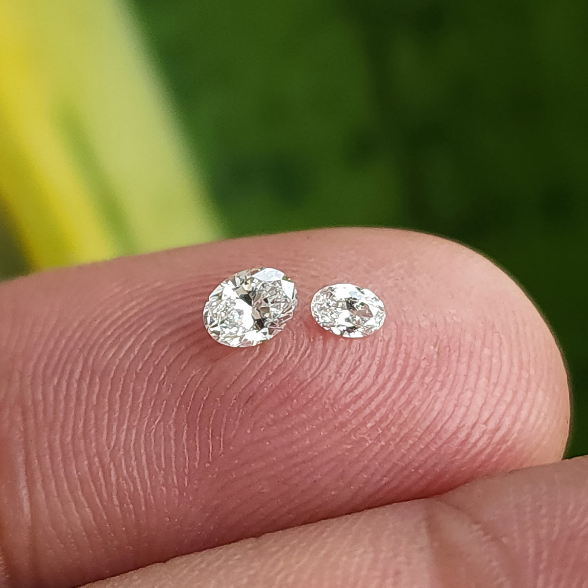 Two oval brilliant diamonds of different sizes resting on a fingertip for size comparison.