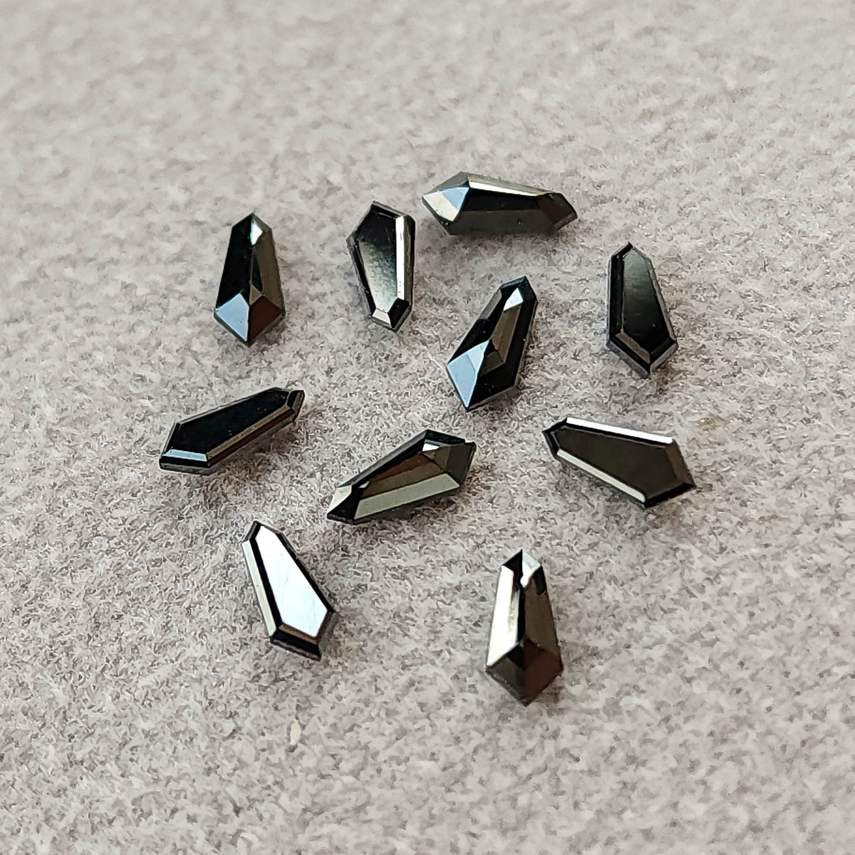Ten black shield-shaped diamonds scattered on gray textured paper, with bright reflections along their edges.