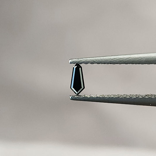 One black shield-cut diamond held face-up in tweezers against a pale gray background, with bright narrow border facets.