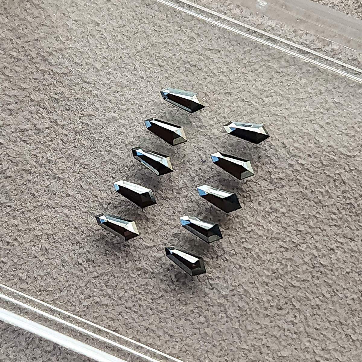 Ten black shield diamonds arranged on gray paper beneath a clear display-box lid, with alternating reflective faces.