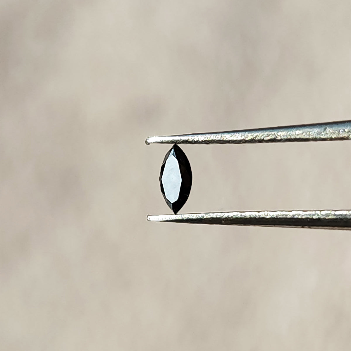 Black marquise diamond in tweezers with a bright reflection across its elongated flat table.