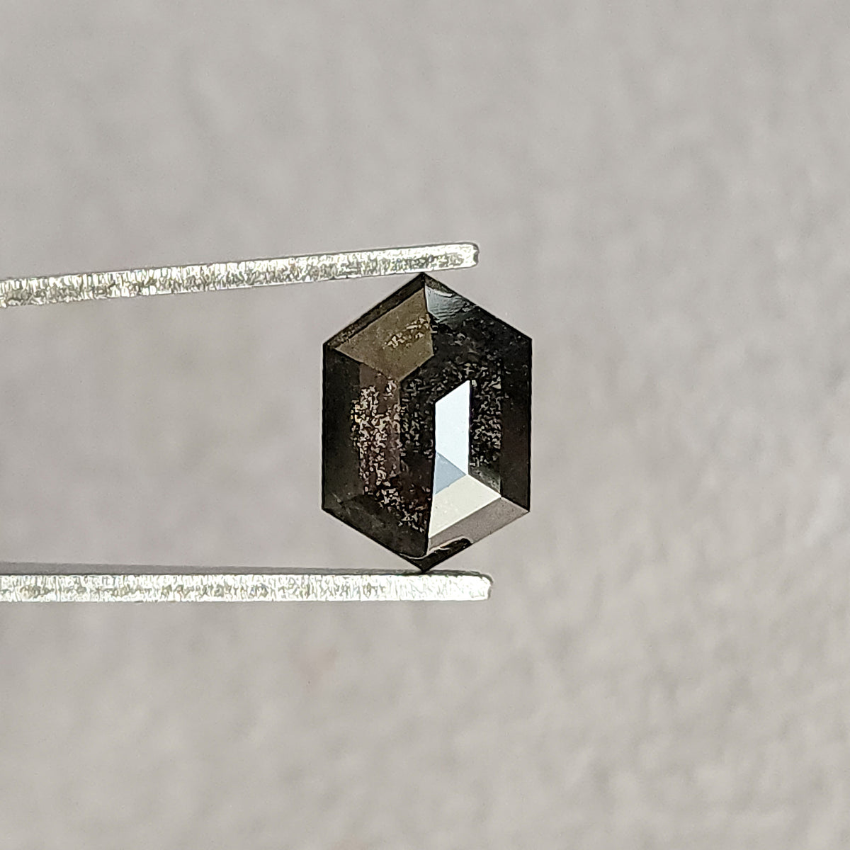 Hexagonal salt and pepper diamond held face-up in tweezers, with a dark included center and bright rose-cut facet reflections against gray.