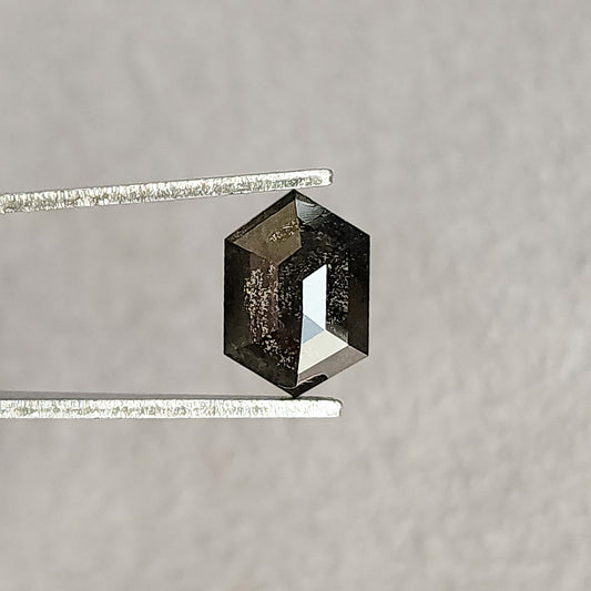 Hexagonal salt and pepper diamond held face-up in tweezers, with a dark included center and bright rose-cut facet reflections against gray.