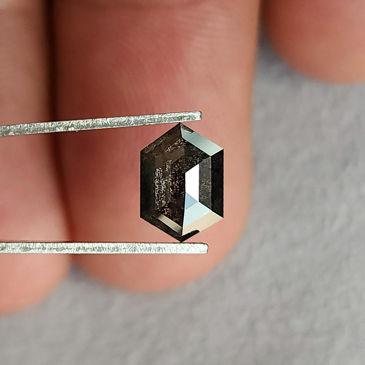 Hexagonal diamond in tweezers with fingers behind it for scale; dark inclusions contrast with bright facets along the right side.