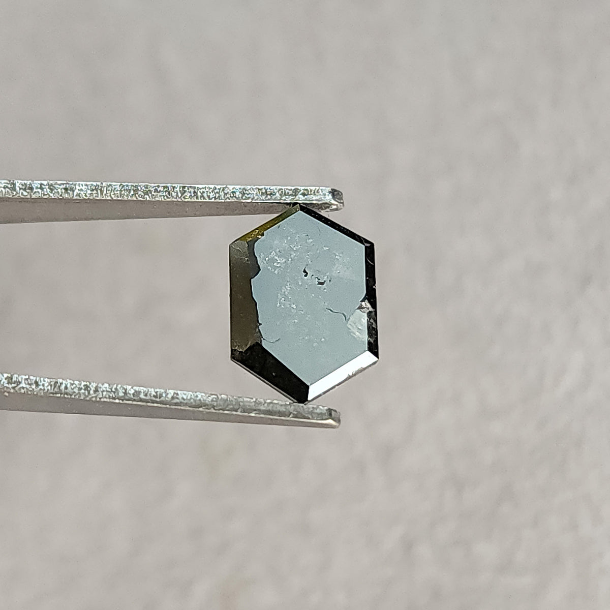 Flat reverse side of the hexagonal diamond in tweezers, with a broad pale reflection and small visible marks against gray.