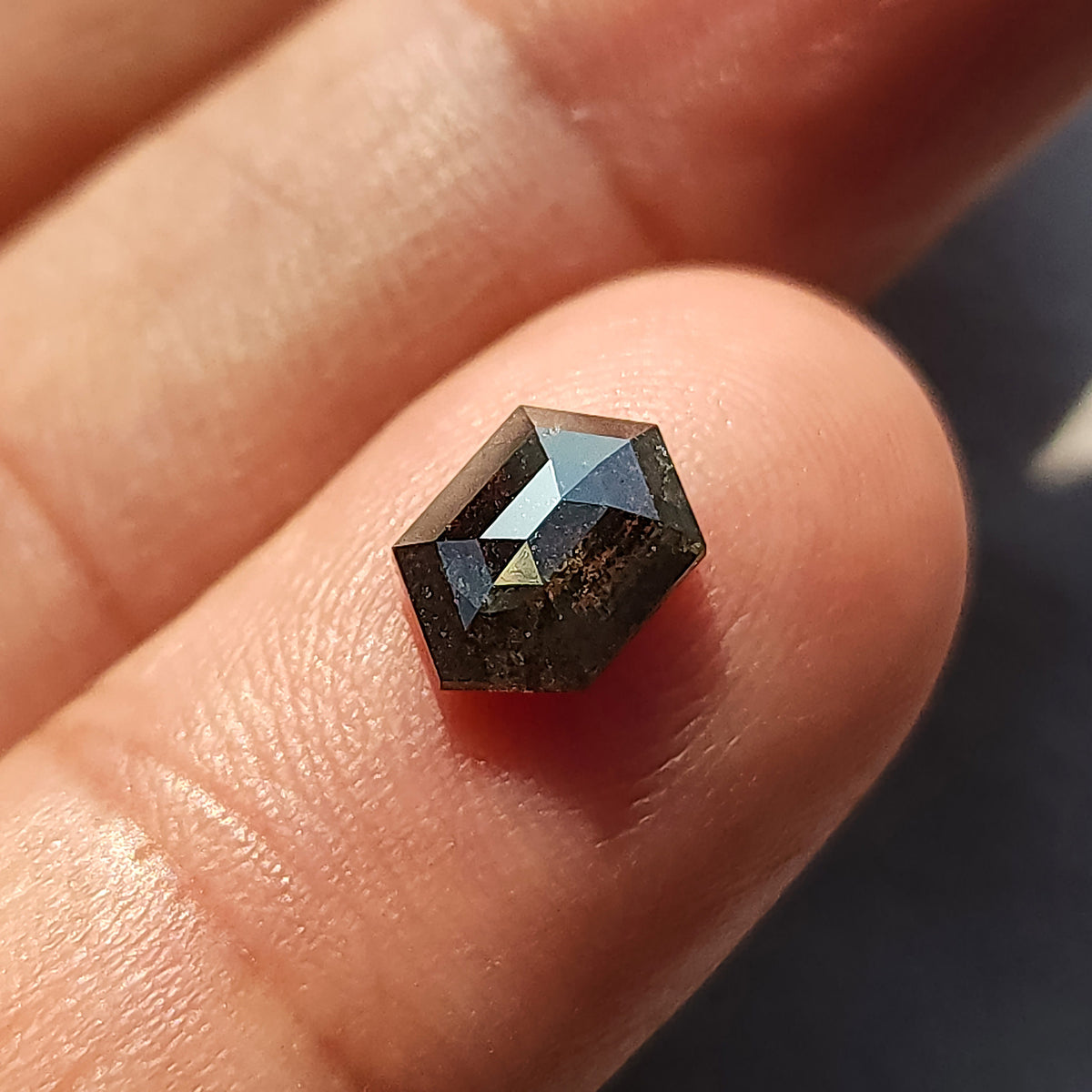 Rotated hexagonal salt and pepper diamond on a fingertip, with pale facet reflections across the upper crown.