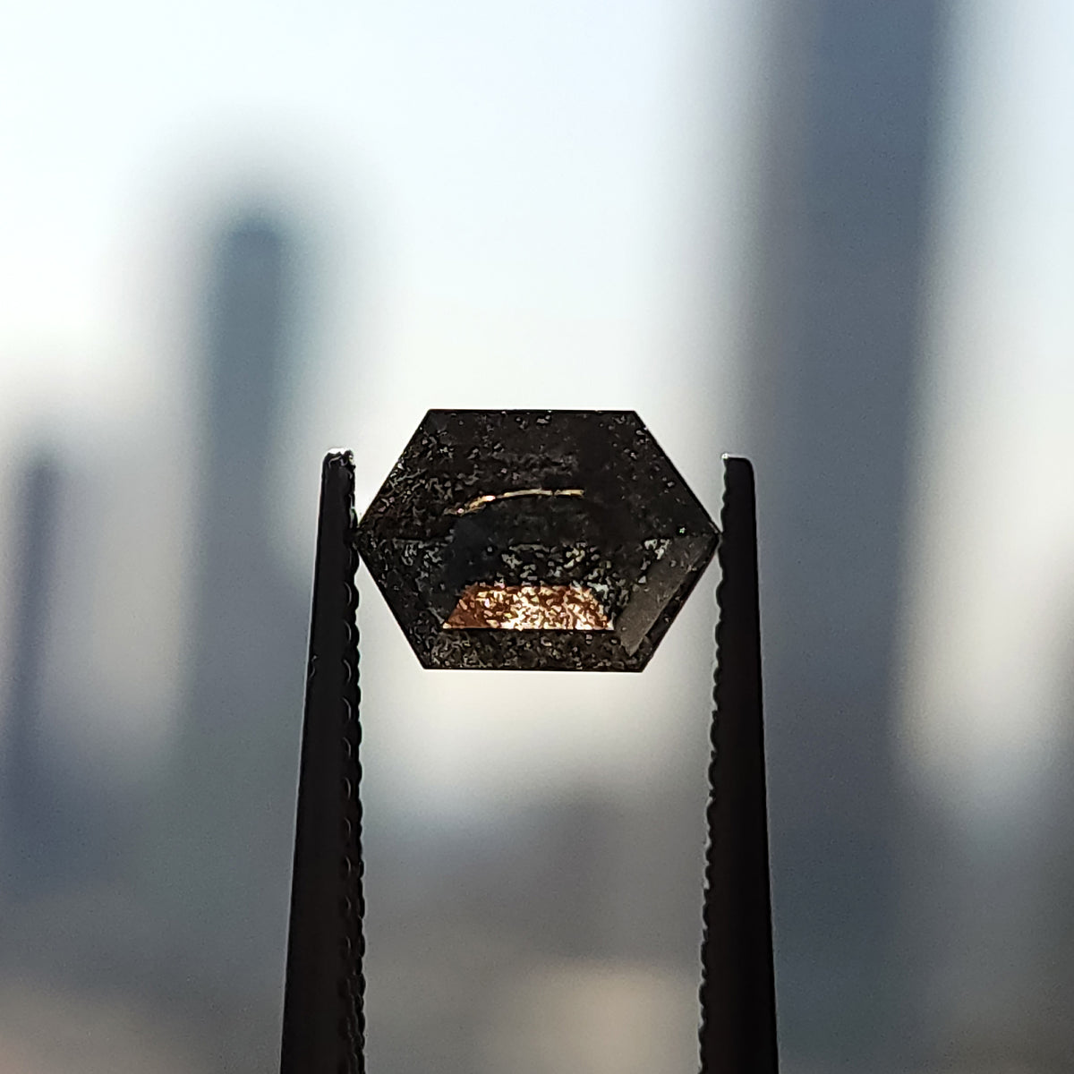 Hexagonal diamond held in upright tweezers against a bright background, showing dark inclusion patterns and a warm patch of transmitted light.