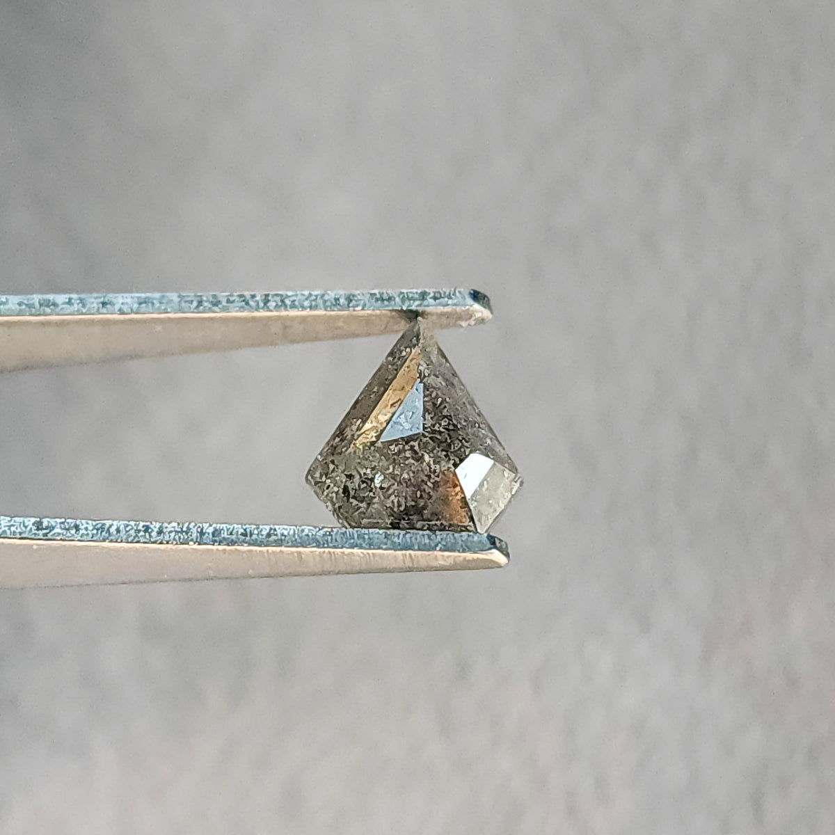 Shield-shaped salt and pepper diamond in tweezers, with pale lower facet reflections and dark inclusions.