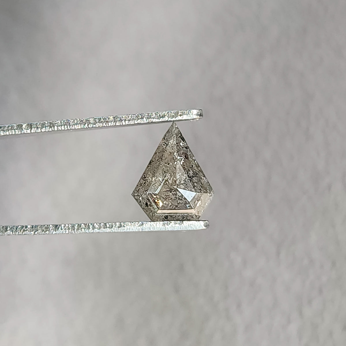 Face-up shield-shaped salt and pepper diamond in tweezers, with a dark included center and bright lower-right facet.